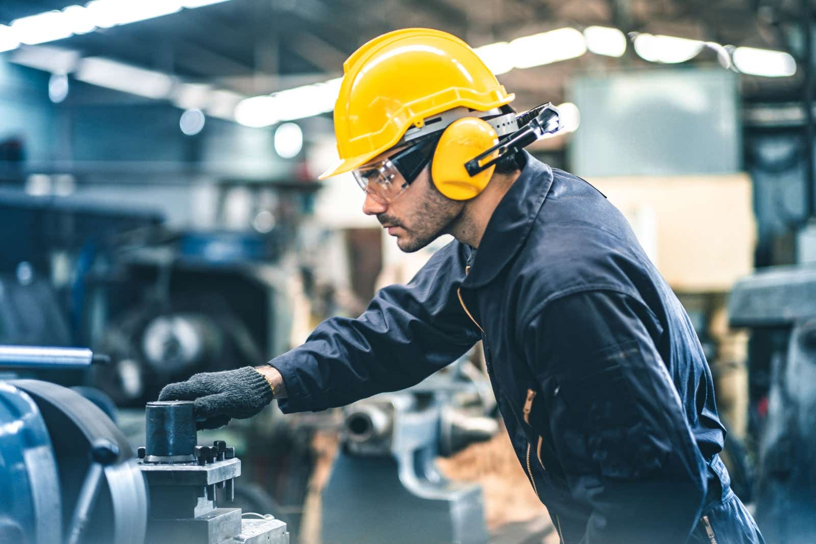 Man in boilersuit, hard hat and ear defenders working in a machine shop.