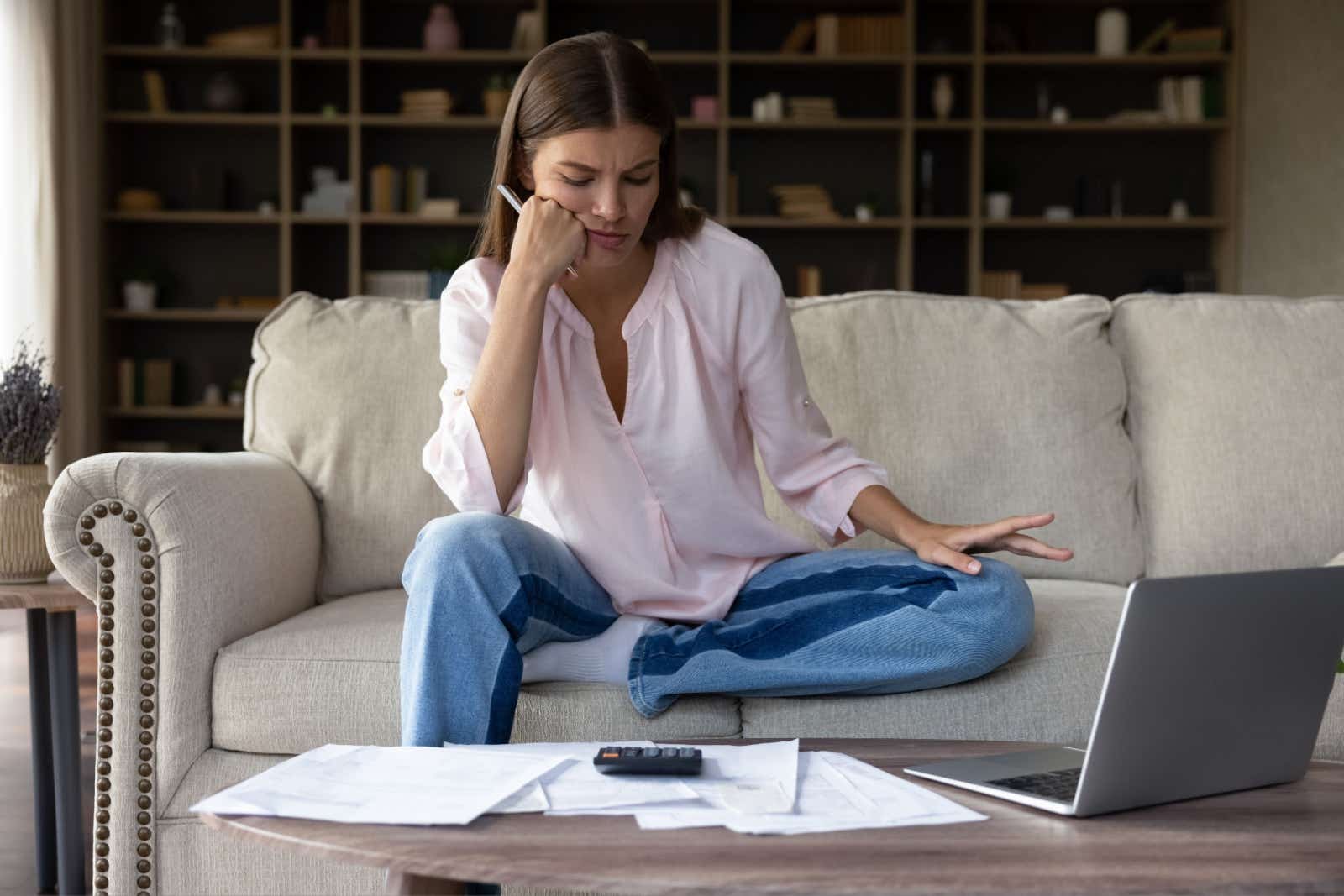 A casually dressed woman seated on a couch looking at paperwork with a calculator. A laptop is off to the side.